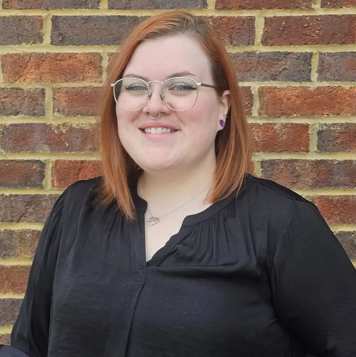 Kelly Whisman, a massage therapist with shoulder-length auburn hair and round glasses, smiling in front of a red brick wall.